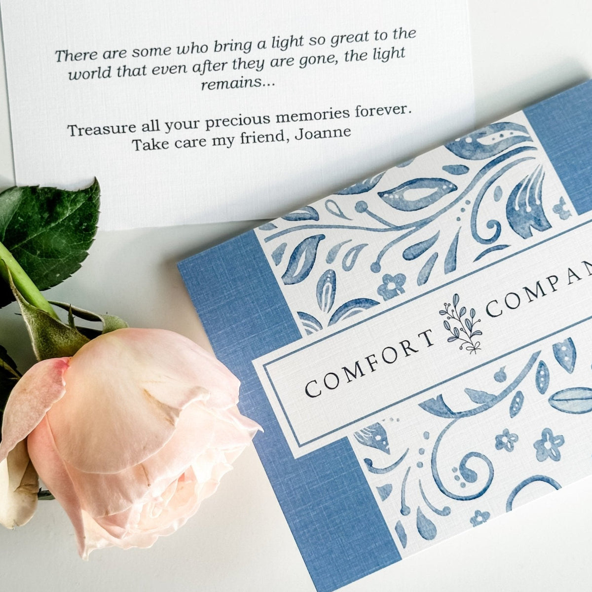 A light pink rose placed beside a blue and white box labeled "COMFORT COMPANY". Above the box is a printed card with a heartfelt message. The scene is set on a white background, emphasizing the stationery and floral arrangement.