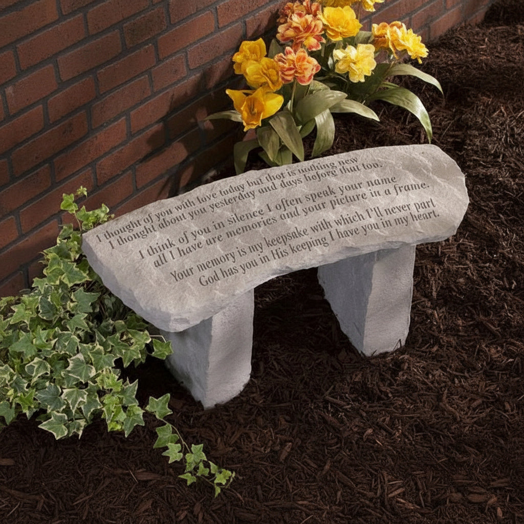 A gray stone memorial bench engraved with a heartfelt message ("I thought of you with love today but that is nothing new, I thought about you yesterday and days before that too. I think of you in silence I often speak your name, all I have are memories and your picture in a frame. Your memory is my keepsake with which I'll never part God has you in His keeping I have you in my heart.")sits in a garden surrounded by flowers and mulch, next to a brick wall.