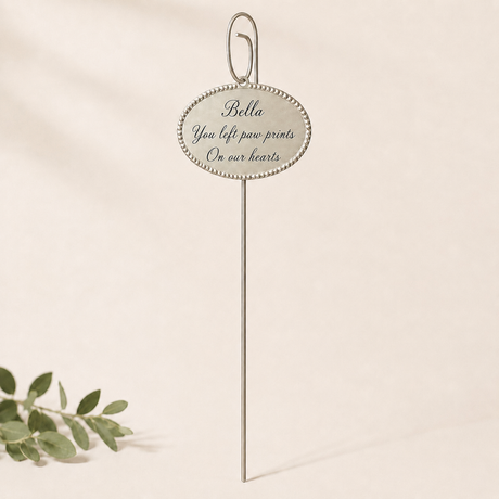 A slender silver memorial pick with an oval beaded plaque reading 'Bella, You left paw prints on our hearts.' It stands centered against a soft cream backdrop, with a small green sprig in the lower left adding a gentle, airy touch.