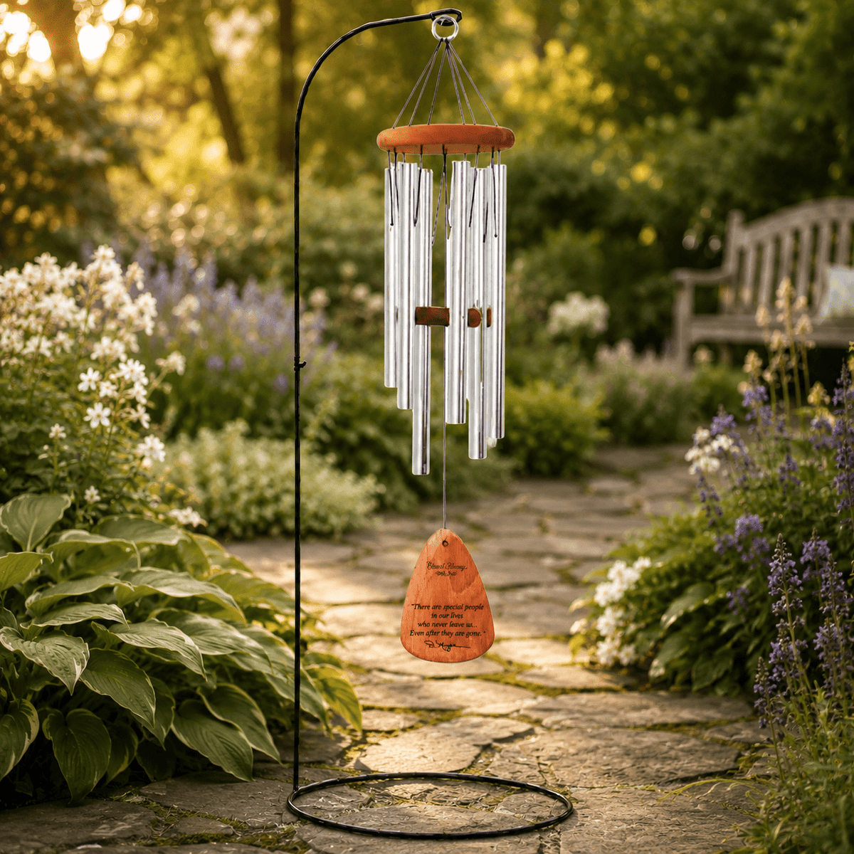 A garden wind chime with silver metal tubes, a warm orange wooden top, and an orange teardrop-shaped hanging weight printed with a poem, "There are special people in our lives who never leave us, Even after they are gone".  Chime is displayed on a black metal stand in a sunlit garden pathway.