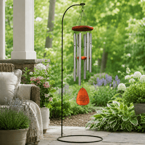 A silver wind chime with an orange top and pendant hangs from a black metal stand on a stone patio. Chime reads, "Your life was a blessing, your memory a treasure...You are loved beyond words and missed beyond measure. Potted flowers, a wicker chair, and lush green garden plants fill the soft daylight scene. 