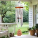 A hanging silver wind chime with an orange top and wooden clapper with wording: Mothers hold their children's hands for a little while but their hearts forever, hanging from the ceiling of a serene outdoor porch.