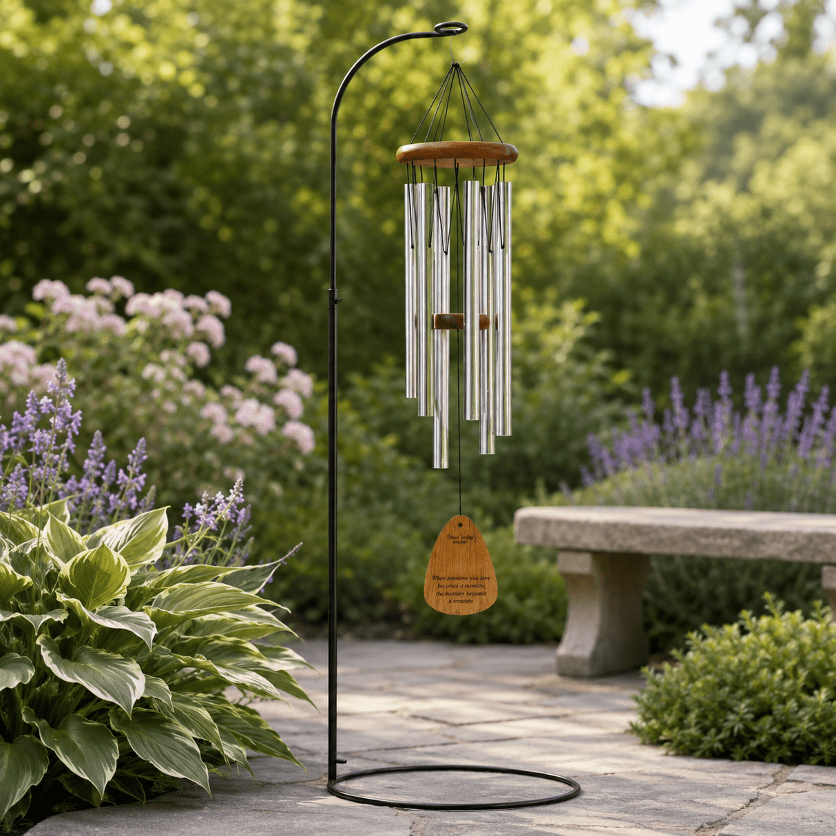 A black metal garden wind chime stands on a circular base, with silver tubes and a small wooden tag which reads, When someone you love becomes a memory, the memory becomes a treasure, hanging beneath a rounded wood top. It is set among lush Hosta, lavender, and a stone bench, with soft green foliage blurred behind.