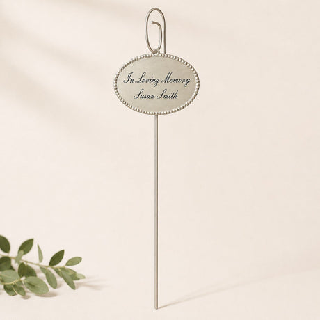 An oval silver memorial sign on a slender metal stake reads “In Loving Memory Susan Smith” in script. Set against a soft cream background with a small leafy sprig in the lower left, it feels delicate and understated.