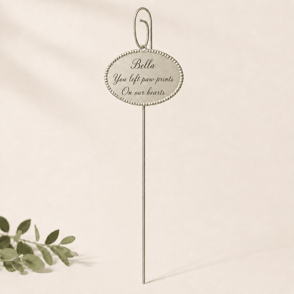 A slender silver memorial pick with an oval beaded plaque reading 'Bella, You left paw prints on our hearts.' It stands centered against a soft cream backdrop, with a small green sprig in the lower left adding a gentle, airy touch.