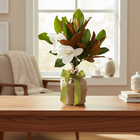 A charming arrangement of magnolia flowers in full bloom, nestled among glossy green leaves, sits elegantly in a natural burlap wrap adorned with a fresh lime green ribbon. The background features a softly lit room with large windows, enhancing the serene atmosphere.