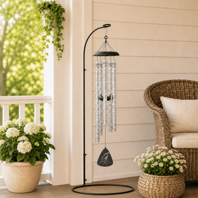 A black metal wind chime with clear tubes hangs from a stand on a patio. Nearby, a wicker chair sits beside potted plants with white flowers and a background of greenery.
