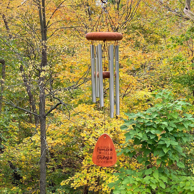 A bronze wind chime with long silver tubes and an orange wooden catcher hangs among dense autumn trees. Set against golden leaves and fresh green foliage, it is centered in the frame under soft daylight, creating a calm woodland scene.